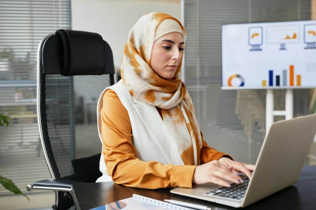 Woman wearing a hijab sitting at a desk and typing on a computer.