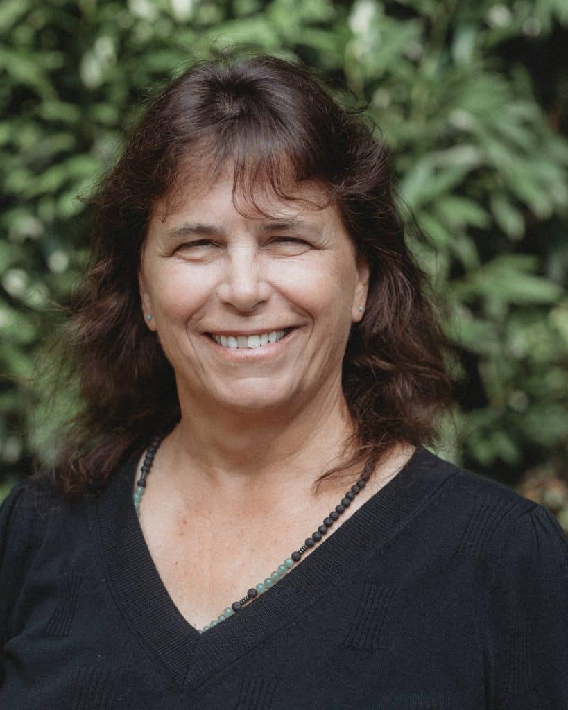 An outdoor headshot of Sue Scavilla wearing a black top and smiling in front of a leafy green background.