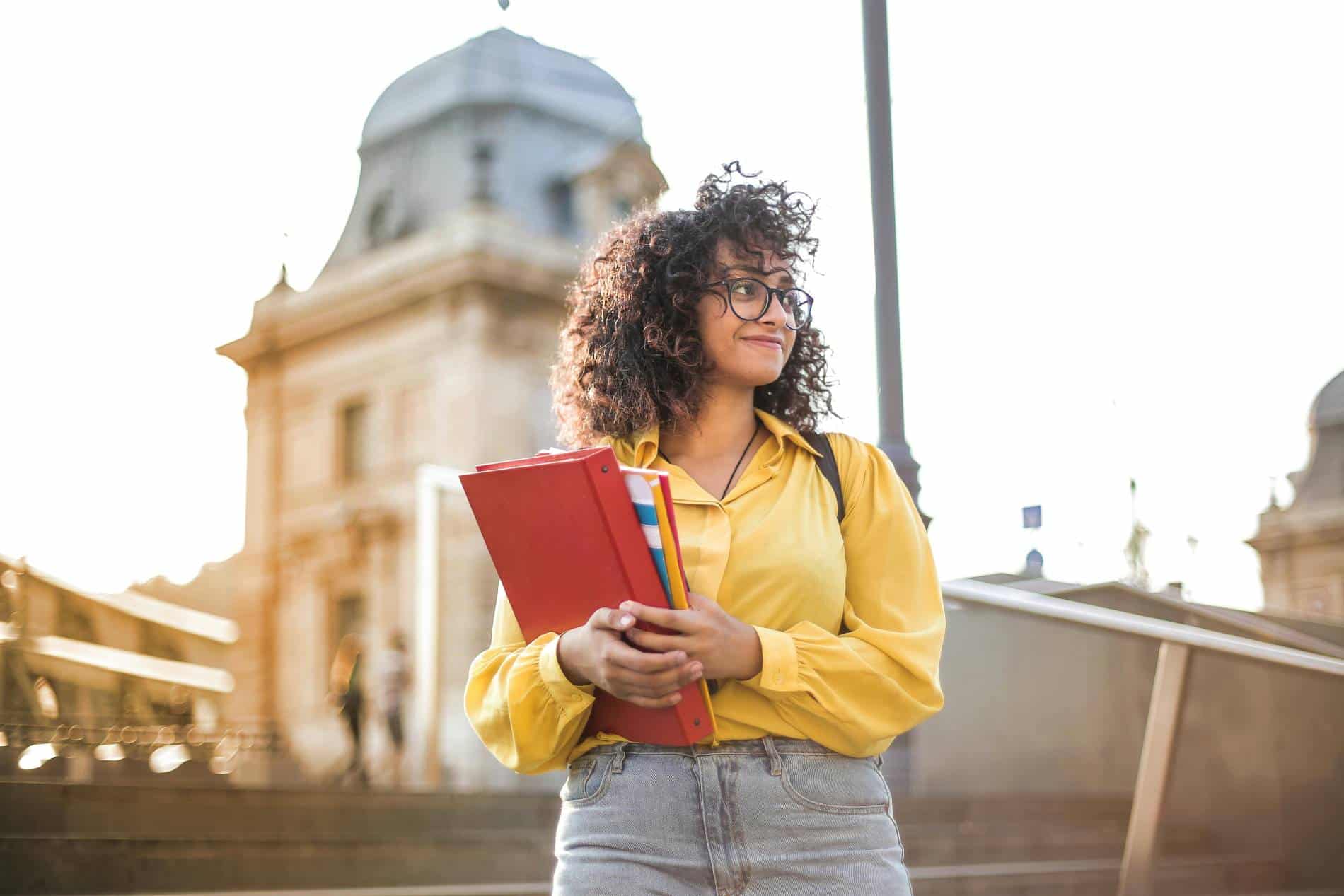 A student holds her books and looks off to the side in front of a stately university building.