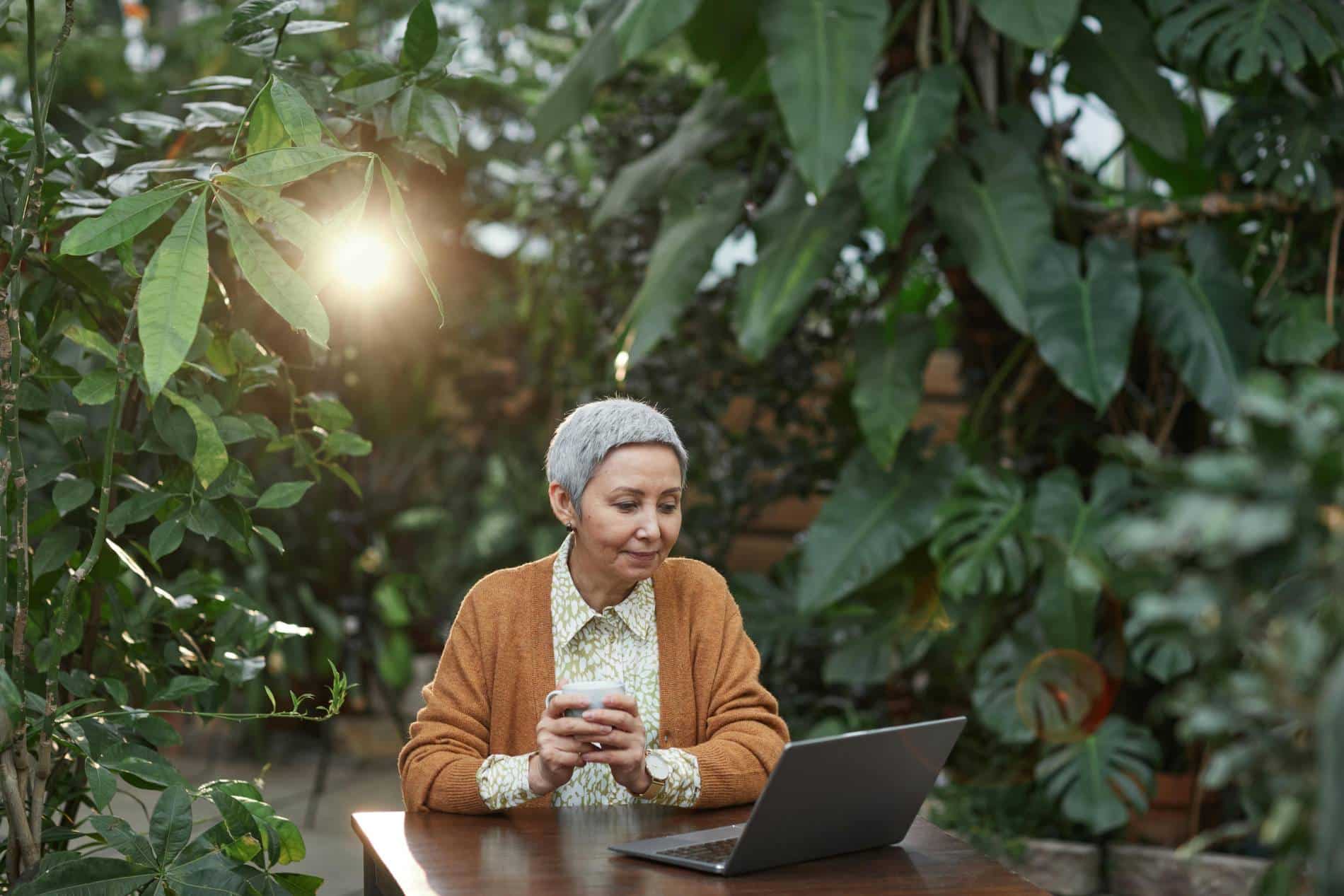 A grey-haired woman sits in a lush,green garden with her laptop and a cup of coffee.