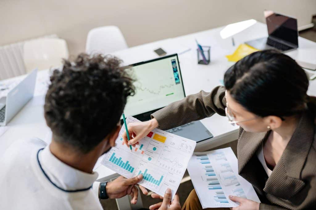 Two people reviewing charts and data at a desk in front of a laptop.